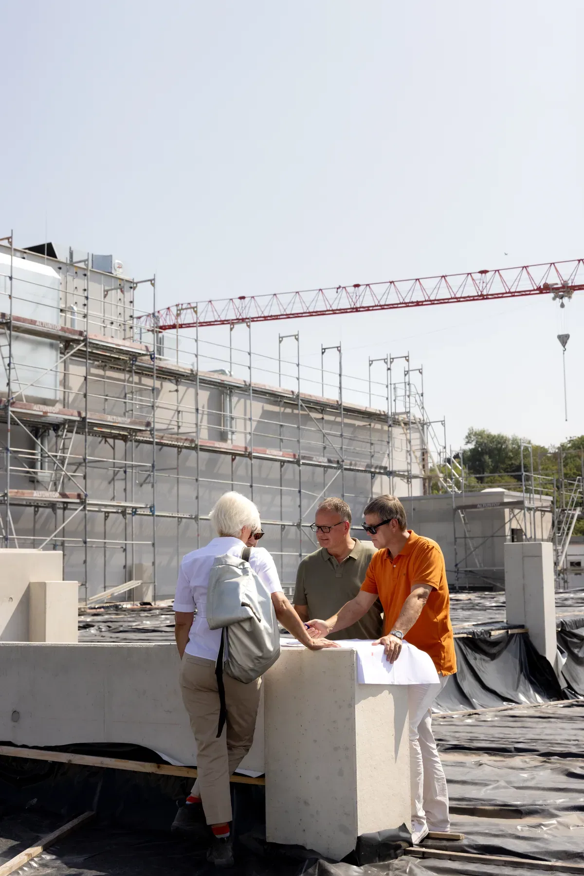 Drei Männer besprechen auf einer Baustelle mit Gerüst und Kran die ausgebreiteten Pläne