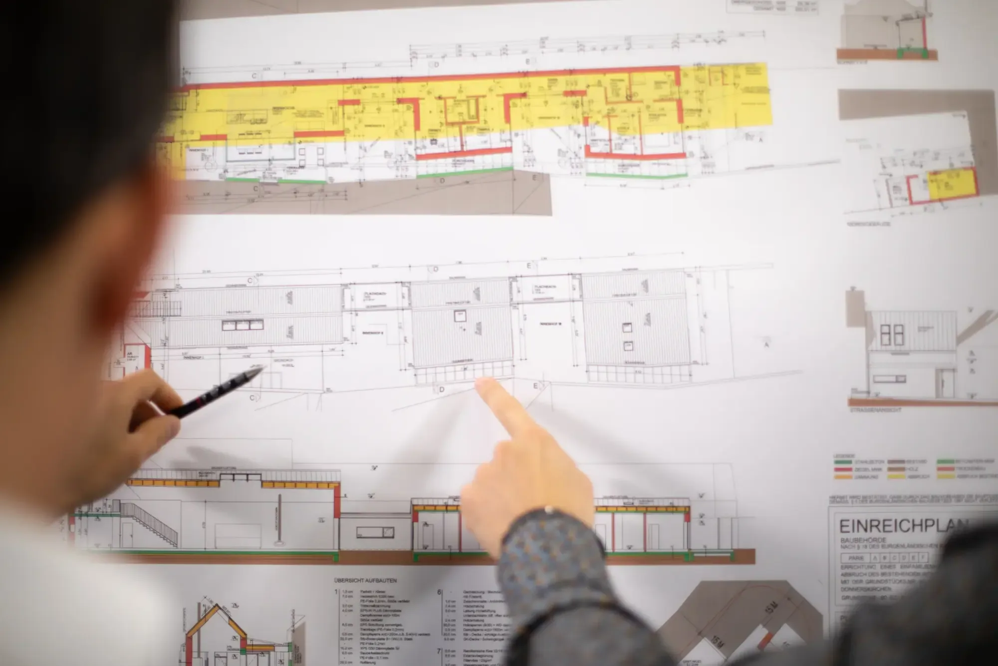 Hände zeigen mit Stift und Finger auf einen Einreichplan an der Wand