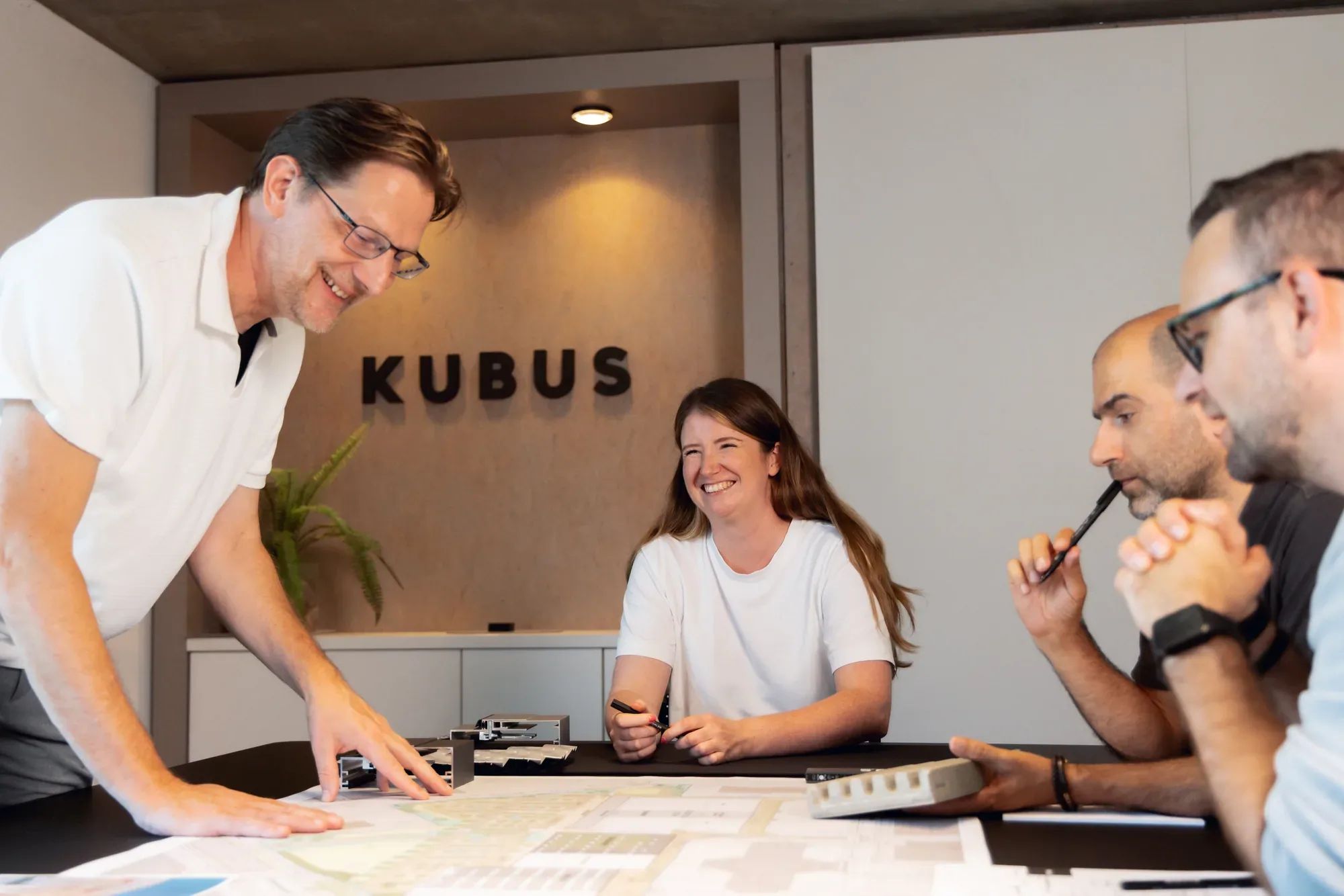 Das Team von KUBUS bespricht lachend Pläne am Tisch vor dem Firmenlogo