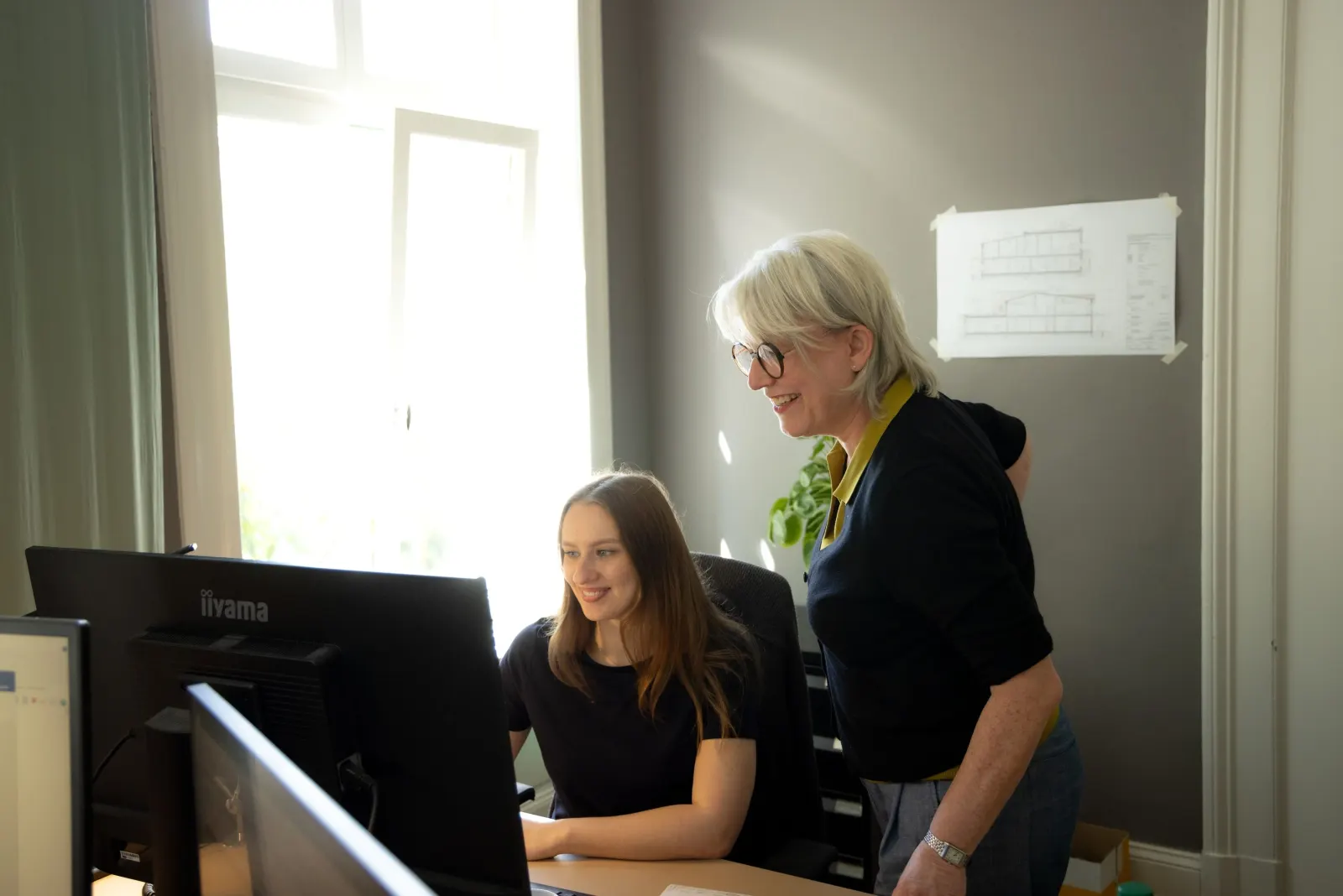 Zwei Frauen arbeiten gemeinsam am Bildschirm in einem hellen Büro, die ältere erklärt der jüngeren etwas