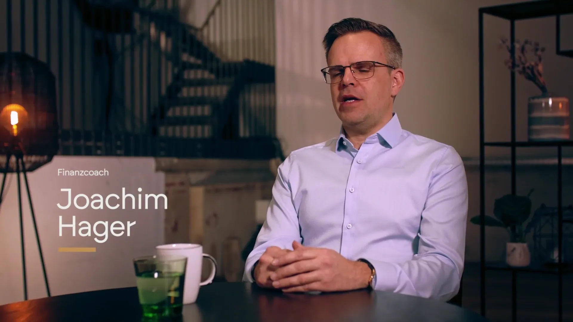 Finanzcoach Joachim Hager im Videointerview am Tisch, daneben ein Aufsteller mit seinem Namen