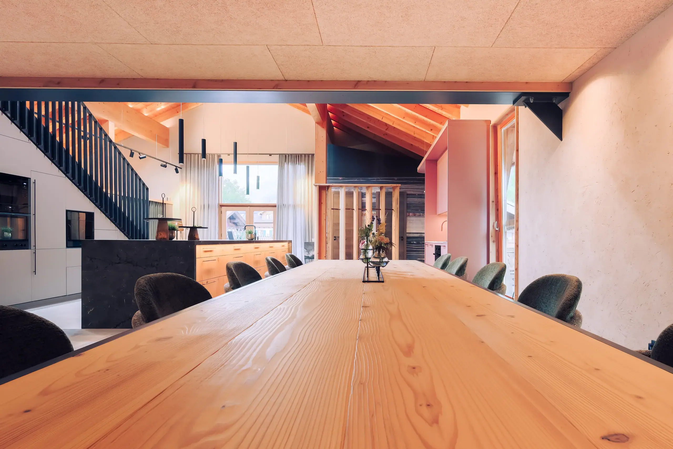 Langer Holztisch mit Stühlen im offenen Wohnraum mit Kücheninsel, Treppe und sichtbarem Dachgebälk