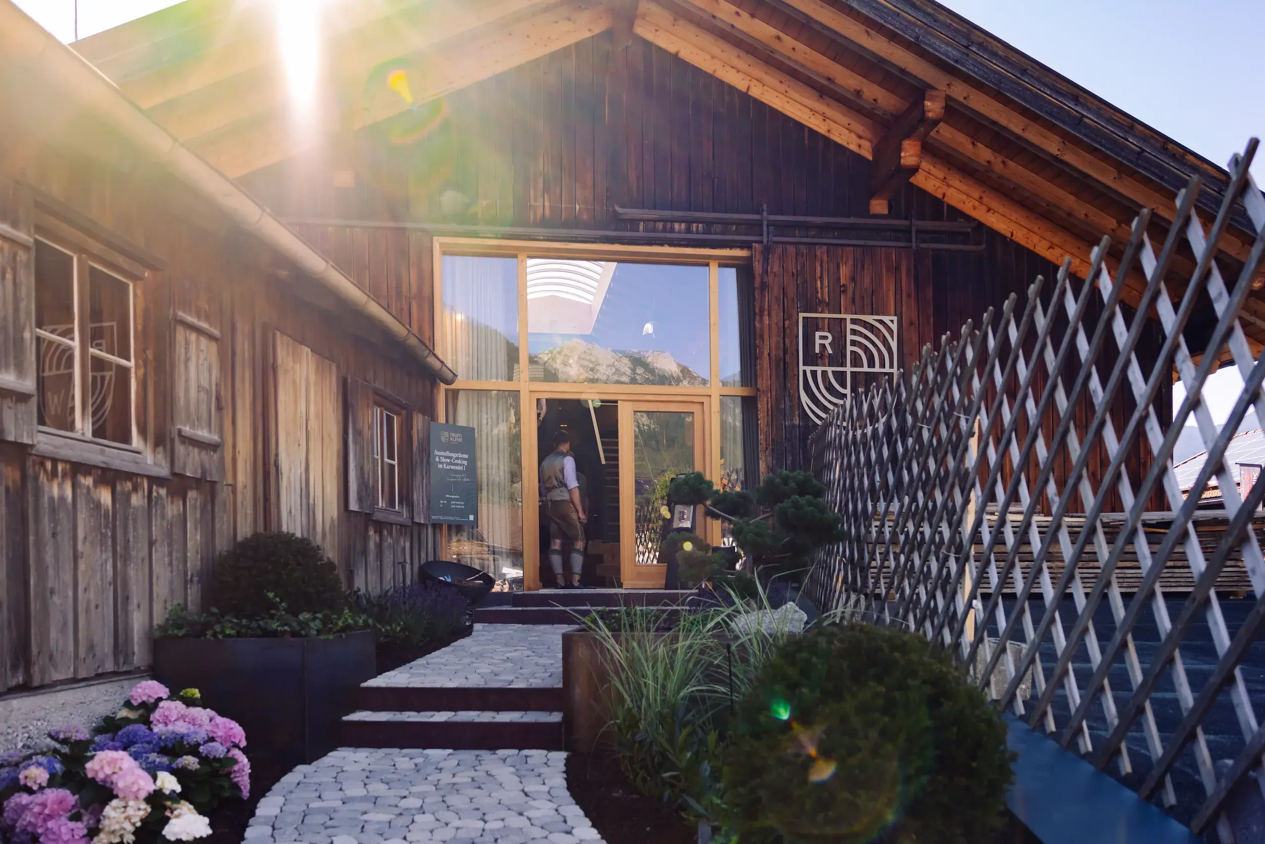 Eingang eines Holzgebäudes mit Glasfront, Natursteinweg und Blumenbeeten bei Sonnenschein, Person im Eingang