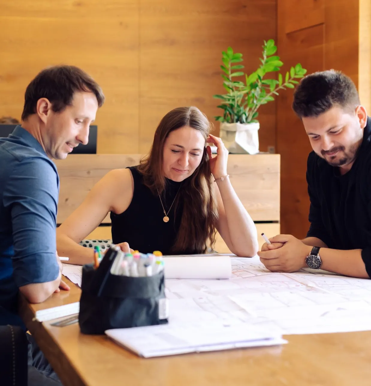 Drei Mitarbeitende besprechen Baupläne an einem Tisch vor einer Holzwand