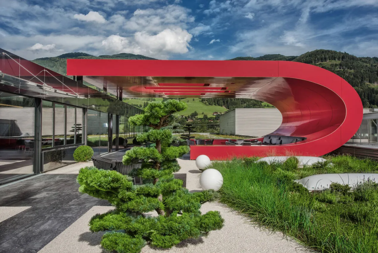 Dachterrasse mit roter, geschwungener Überdachung, Formschnitt-Kiefer und Gräsern vor Bergkulisse
