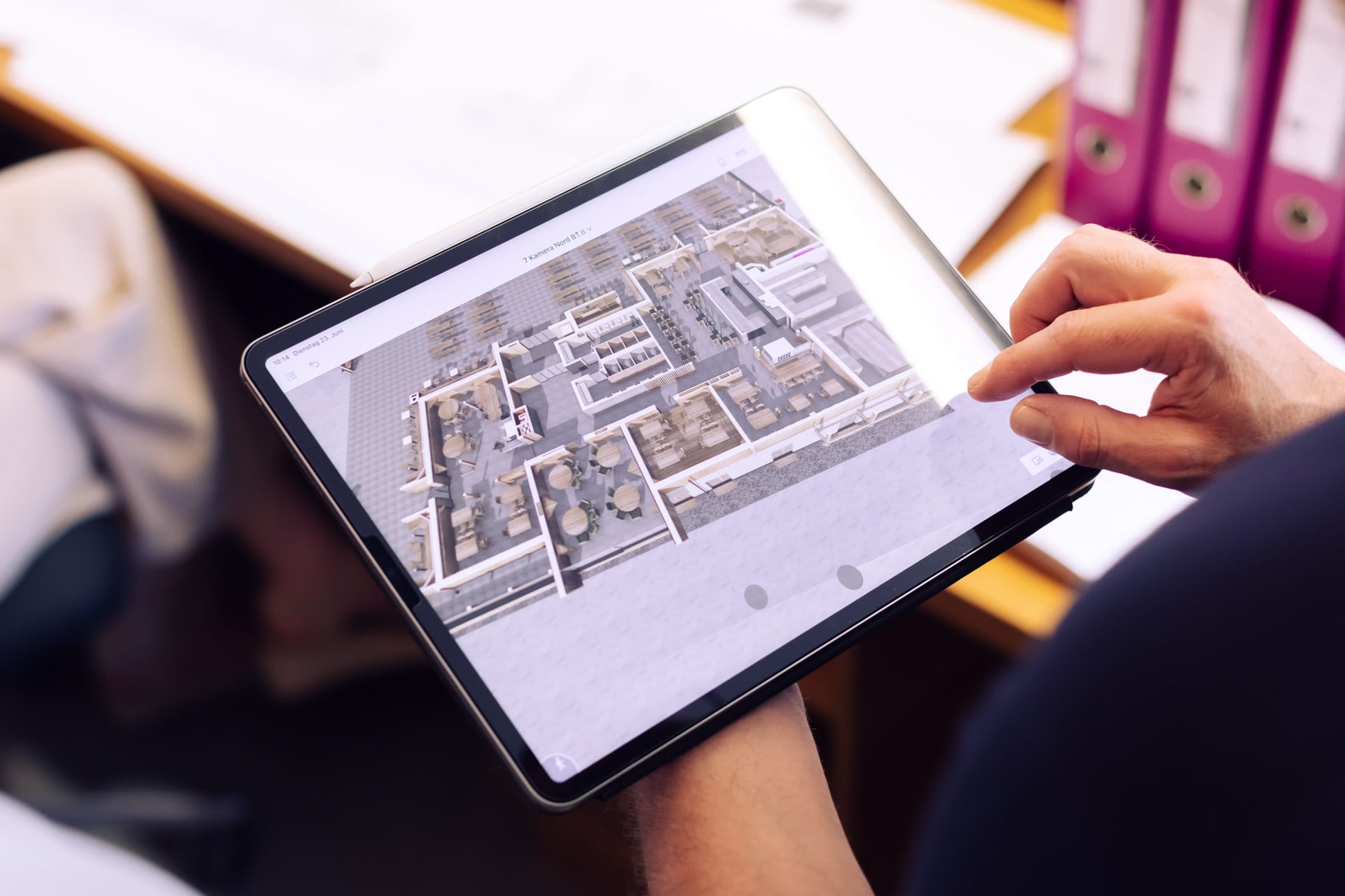 Hand hält ein Tablet mit dem 3D-Modell eines Gebäudes über ausgebreiteten Plänen